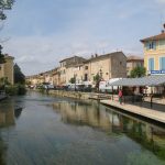 Un canal paisible traversant une ville avec une promenade bordée d'arbres et des bâtiments colorés. Des stands de vendeurs et un bâtiment de la "Police Municipale" sont visibles.