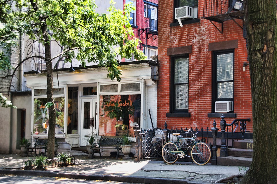 Vue sur la rue d'un café-terrasse pittoresque avec un extérieur blanc à côté d'un bâtiment en brique. Deux vélos sont garés devant et il y a des arbres et des plantes le long du trottoir.